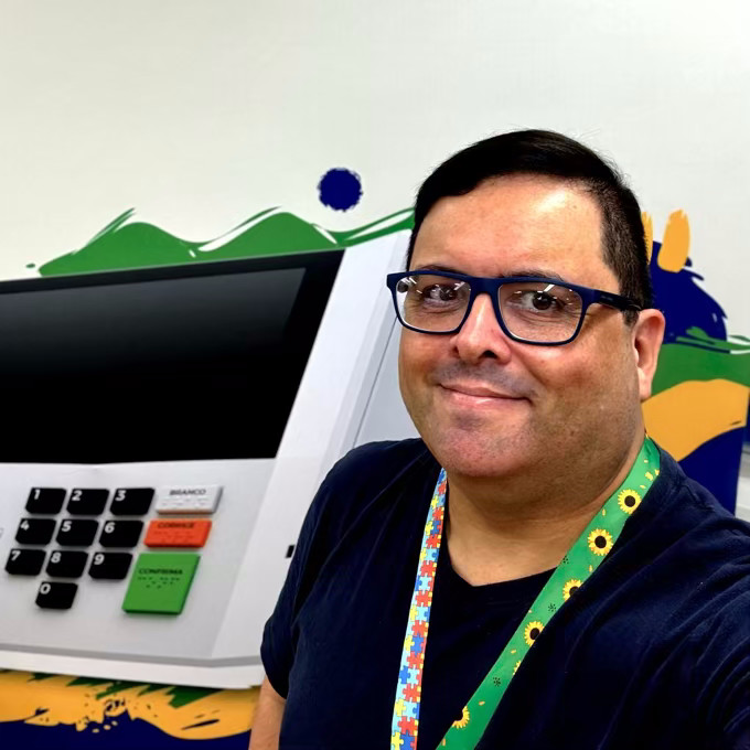 Foto minha, olhando pra câmera, de camiseta azul e usando cordão de crachá com os símbolos do autismo e do girassol, junto a um painel mostrando a imagem de uma urna eletrônica modelo 2023