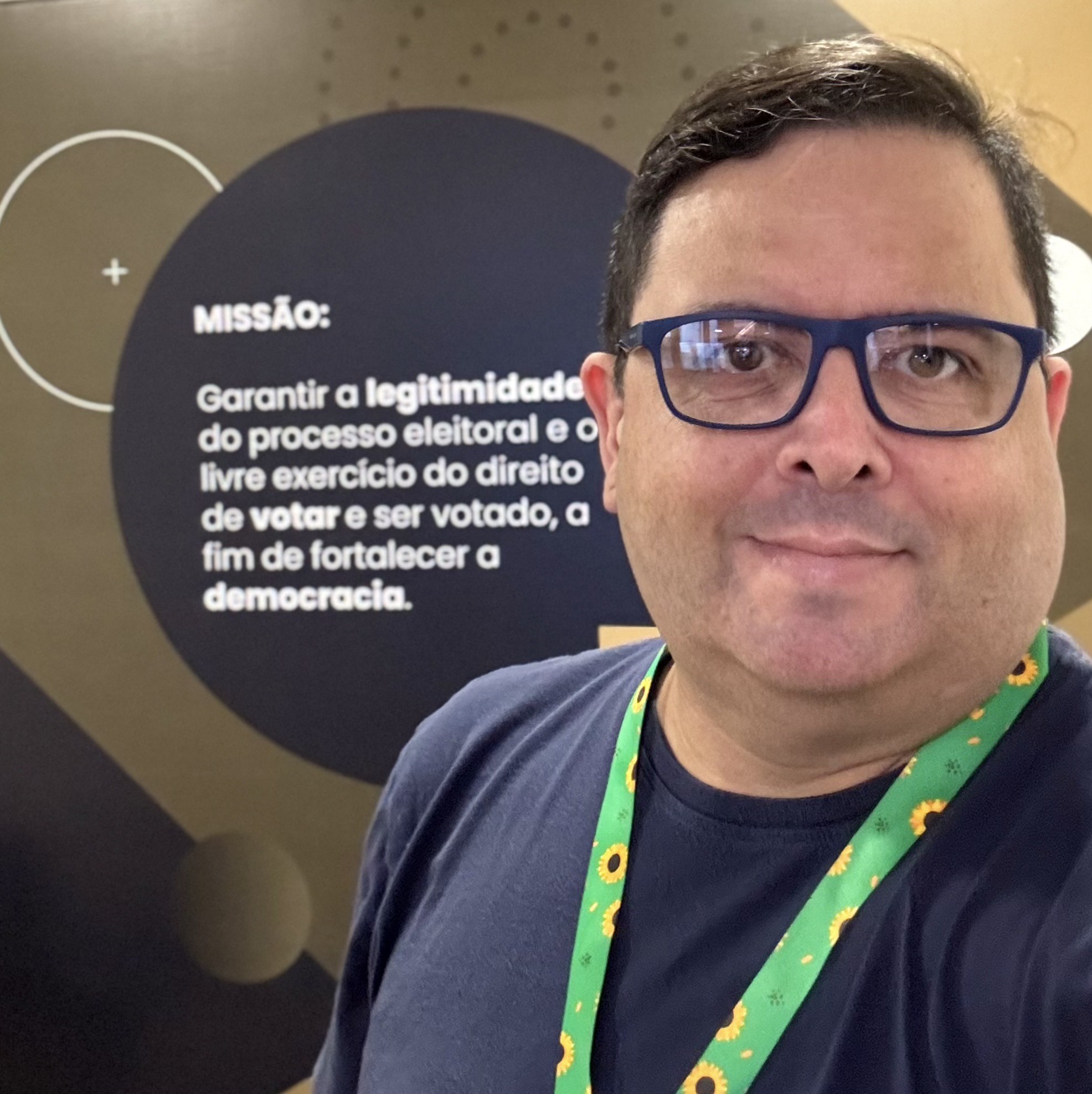 Selfie minha de camiseta azul e cordão de girassol, em frente a um painel onde se lê:<br><br>MISSÃO: Garantir a legitimidade do processo eleitoral e o livre exercício do direito de votar e ser votado, a fim de fortalecer a democracia.
