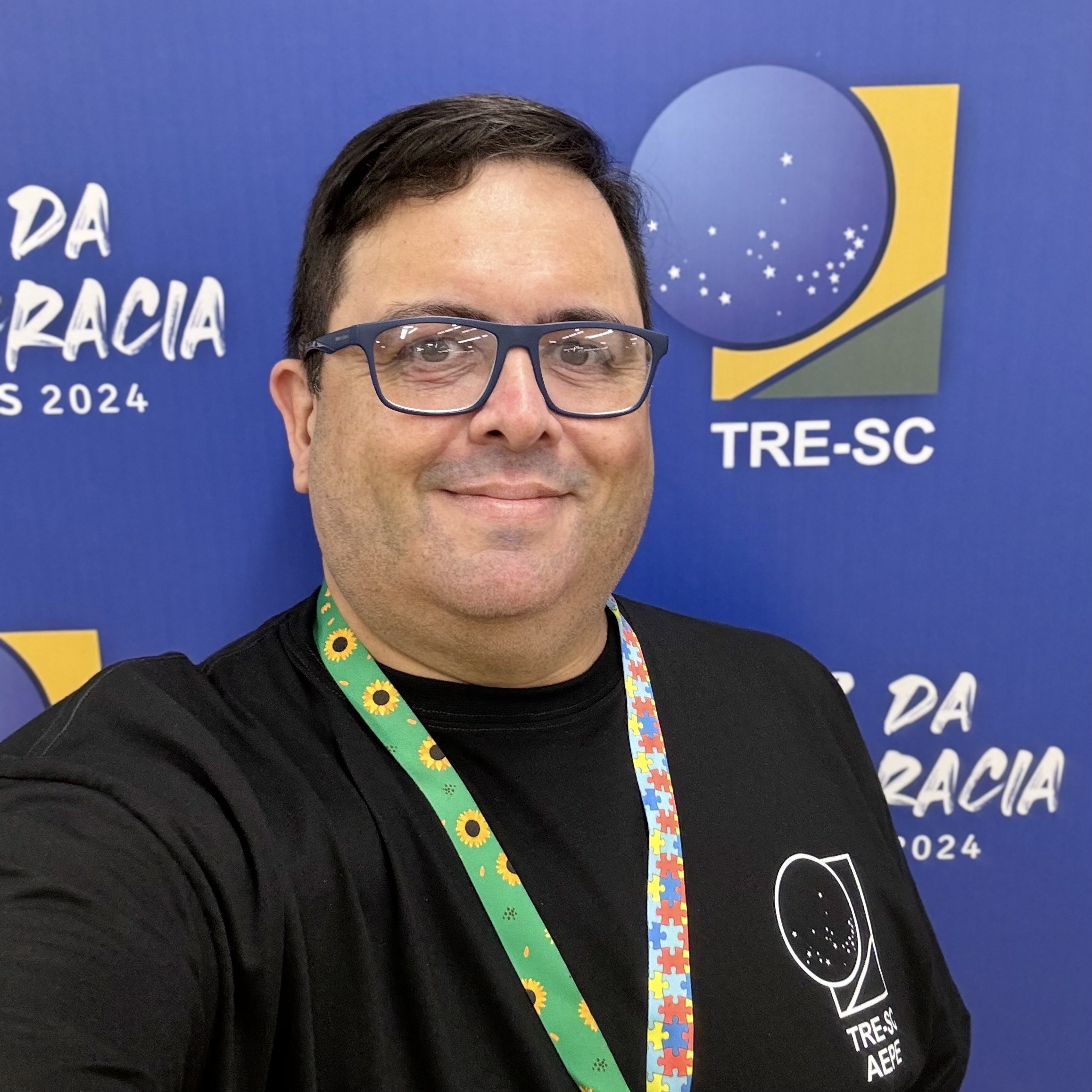 Foto minha com cordão com símbolos de girassol e de quebra-cabeças e camiseta preta, em frente a um banner das Eleições 2024 em SC