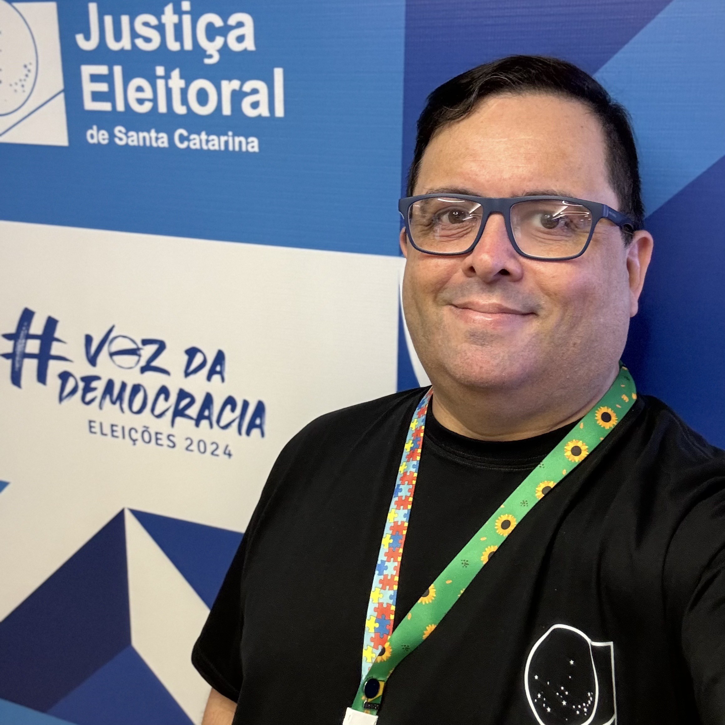 Foto minha de camiseta preta e cordão de crachá com o símbolo do autismo, em frente a um banner com o logo das Eleições 2024