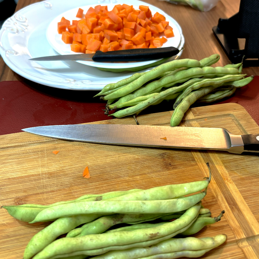 Um maço de vagens, junto a uma faca, na tábua de corte e ao lado de um prato contendo cenoura cortada em cubos