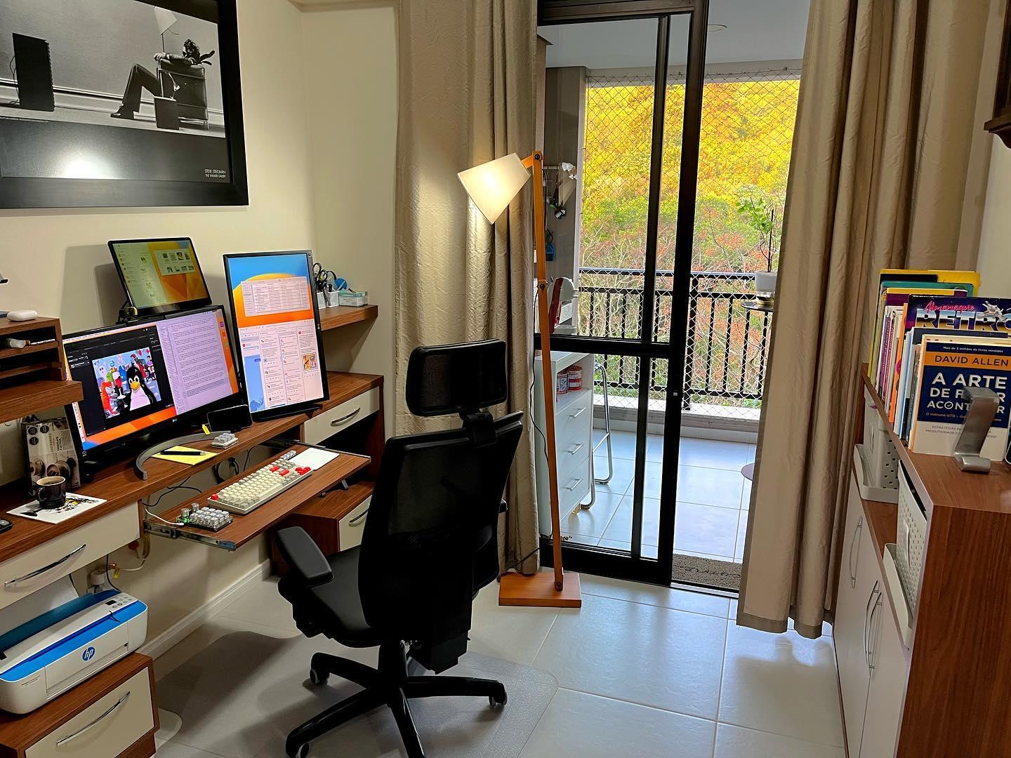 Foto de escritório doméstico com escrivaninha estreita e larga, prateleiras ao redor dos monitores, com gavetas abaixo do tampo e simétricas a elas, e espaço para impressora (e armazenamento dos seus suprimentos) abaixo delas. A estante com material de referência e espaço para guardar os materiais de escritório fica atrás, acessível girando a cadeira.