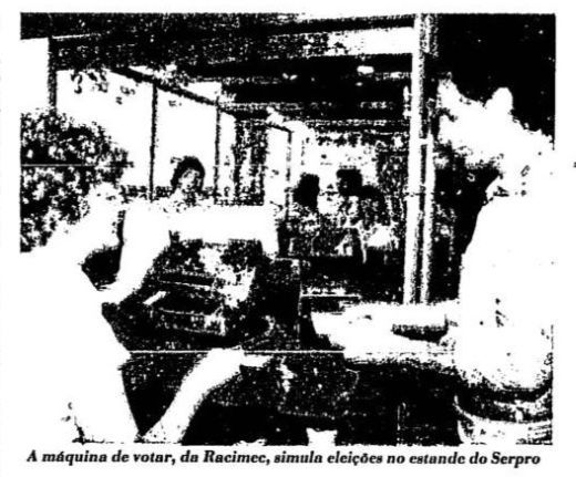 Foto com baixa visibilidade, de jornal da época, mostra duas pessoas em frente a um equipamento, e a legenda: A máquina de votar, da Racimec, simula eleições no estande do Serpro.