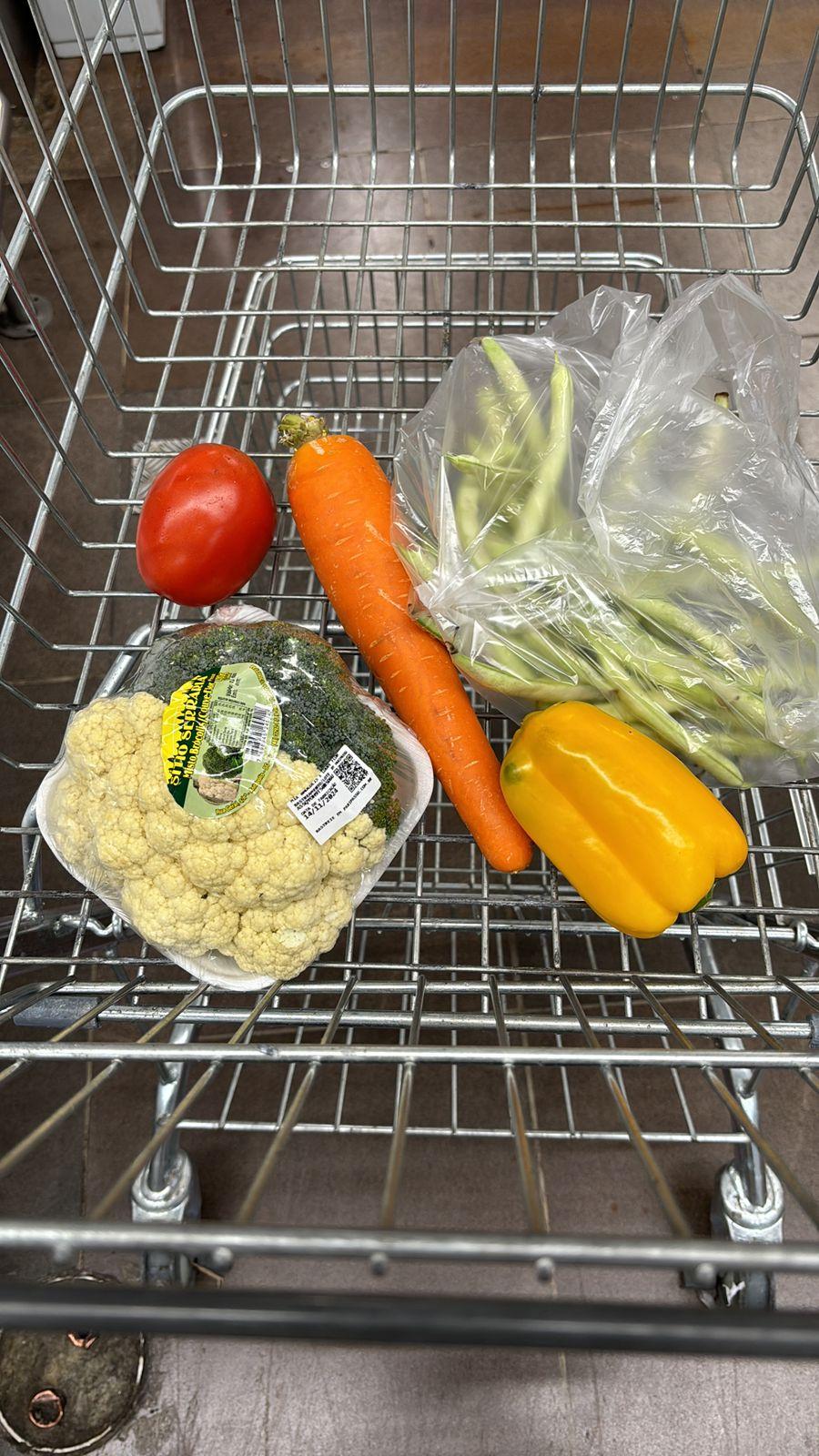 Foto de um carrinho de supermercado contendo uma cenoura, um tomate, um pimentão amarelo, várias vagens, meio brócolis e meia couve-flor