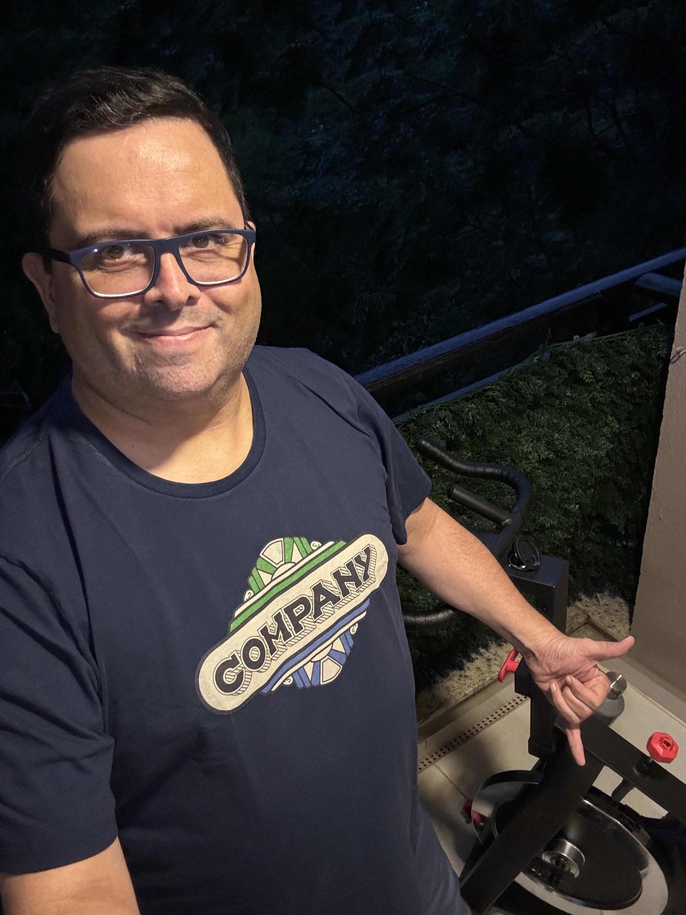 Foto minha com camiseta em estampa vintage da Company, olhando para a câmera e fazendo sinal de hang loose com a mão esquerda, que está próxima ao guidão da bicicleta.