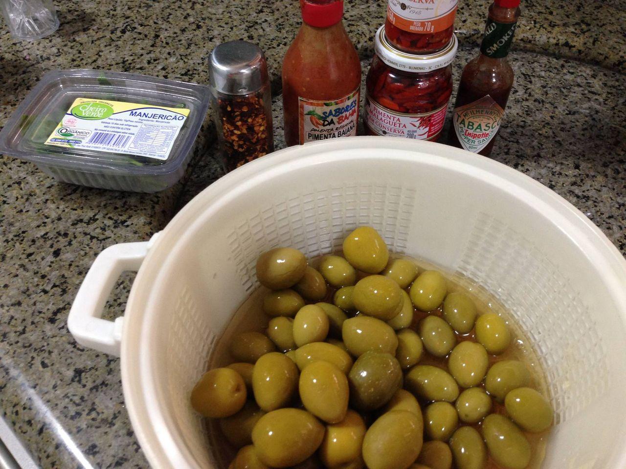 Uma bacia com azeitonas em conserva de vários tamanho, misturadas entre si. Ao fundo, potes de folhas de manjericão, pimenta calabresa, molho de pimenta “sabores da bahia”, molho tabasco chipotle, e um vidro de pimenta malagueta em conserva.