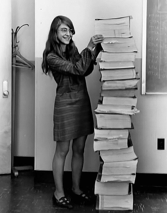Margaret Hamilton sorri junto à pilha de 17 volumes de código-fonte impresso, da programação do computador de bordo da Apollo 11.