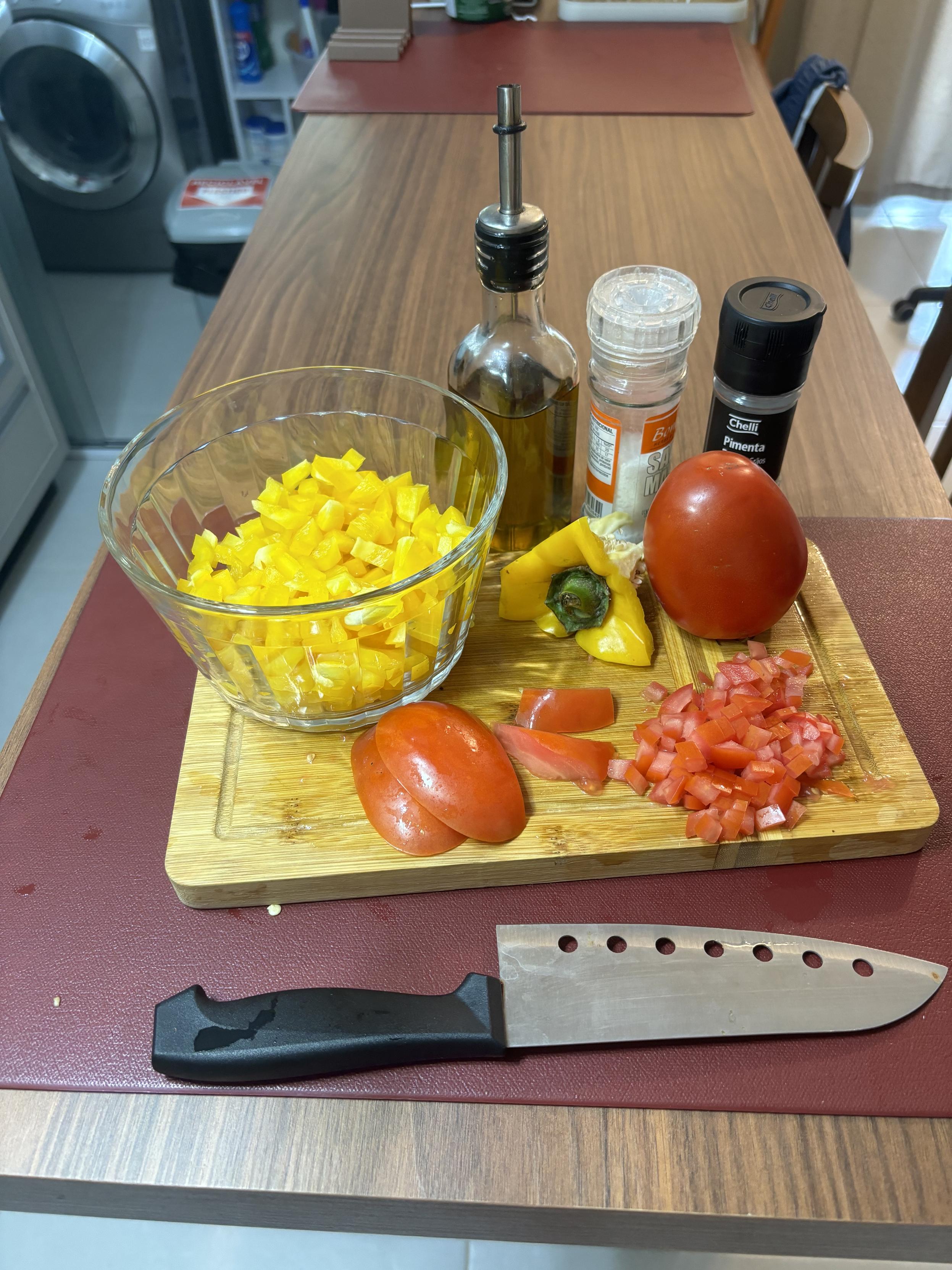 Numa tábua de corte há um pimentão amarelo já cortado em cubinhos, um tomate já parcialmente cortado, e outro tomate ainda inteiro. Ao fundo se vê uma garrafa de azeite, e 2 temperos. 