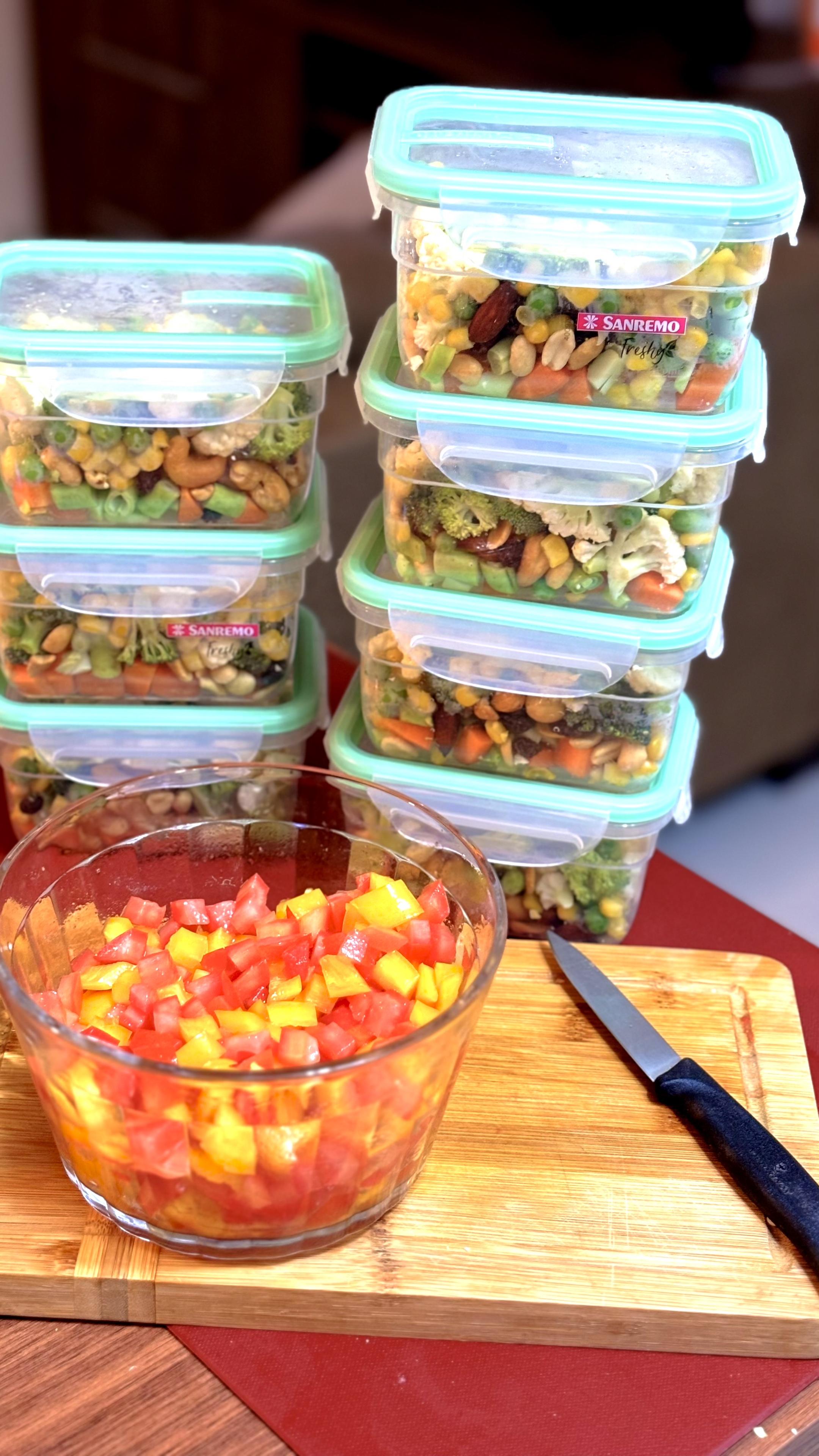 Em primeiro plano uma tigela de vidro contendo um tempero feito com tomate e pimentão picados. Ao fundo, 7 embalagens individuais picadas contendo legumes picados e castanhas. 