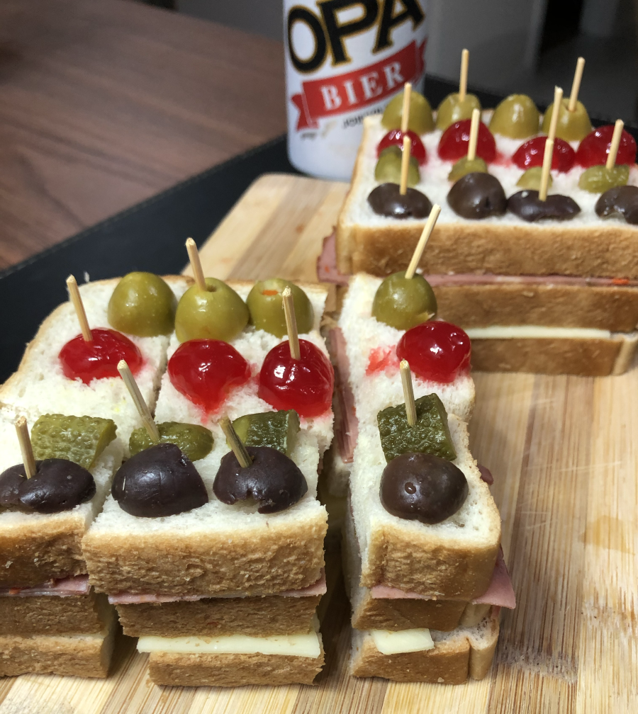 Dois sanduíches, cada um feito com 3 fatias de pão, com uma camada de queijo e uma camada de embutidos. Eles estão decorados com azeitonas de duas cores, cereja e pepino em, conserva. Um deles está cortado em formato petisco, em cubinhos. Ao fundo, uma lata de cerveja marca Opa.