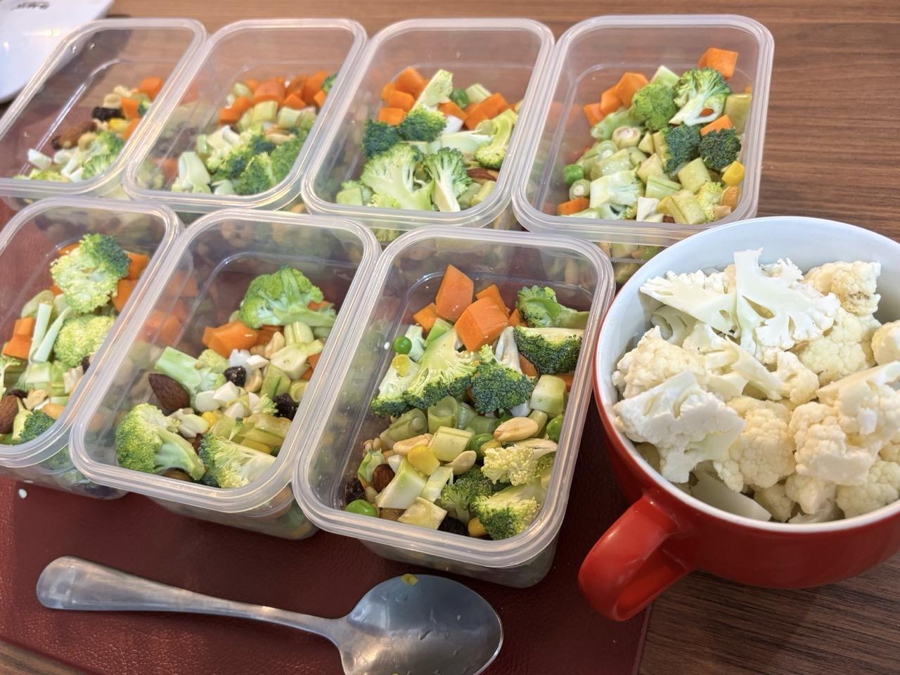 7 potes herméticos, ainda destampados, contendo uma mistura de legumes picados (cenoura, brócolis, couve-flor, vagem), milho, ervilha e sementes. Ao lado deles, uma tigela com alça, contendo couve-flor picada, que ainda será adicionada aos potes.