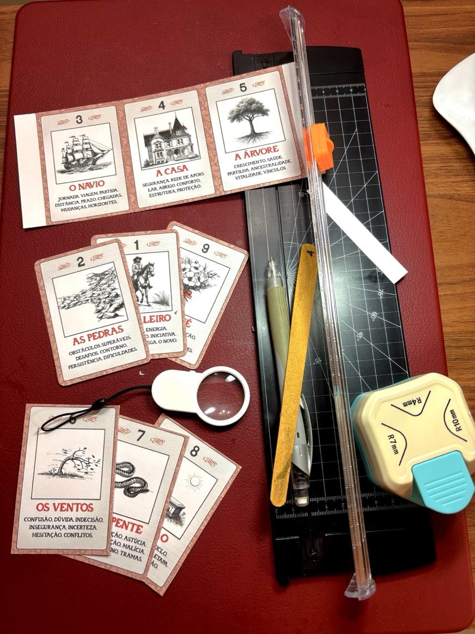 Foto da superfície da minha mesa de trabalho, mostrando as 9 cartas em processo de produção conforme descrito no post, e o material de trabalho: uma mini-guilhotina para papel A4, um cortador de cantos arredondados, lupa, lapiseira e lixa de unha.