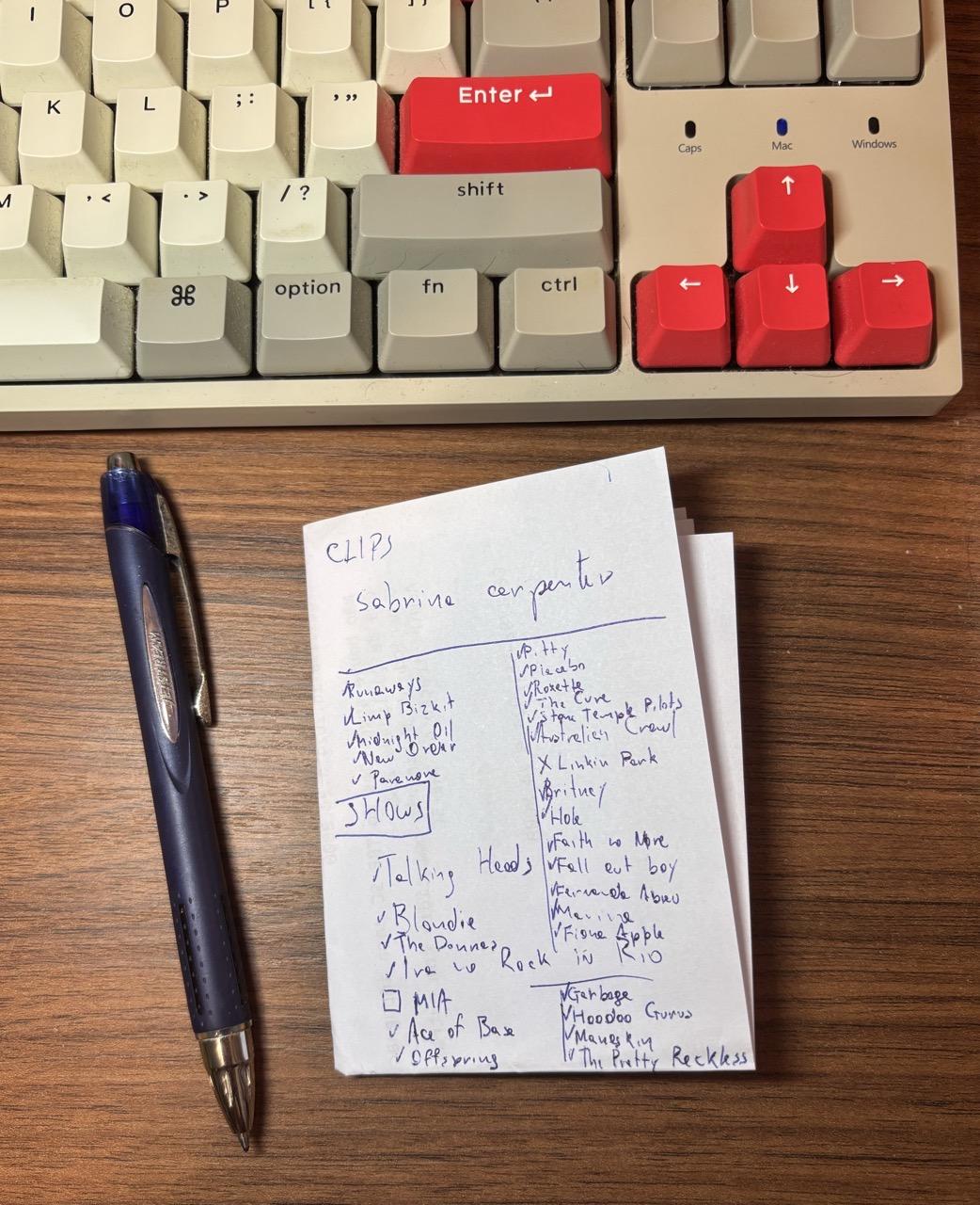 Foto de um papel na minha escrivaninha, aonde estão escritos com a caneta (que está ao lado do papel) os nomes de 29 artistas ou bandas, incluindo Talking Heads, Fernanda Abreu, Blondie, Marina Lima, Maneskin, Sabrina Carpenter, Midnight Oil, Britney e muito mais. 