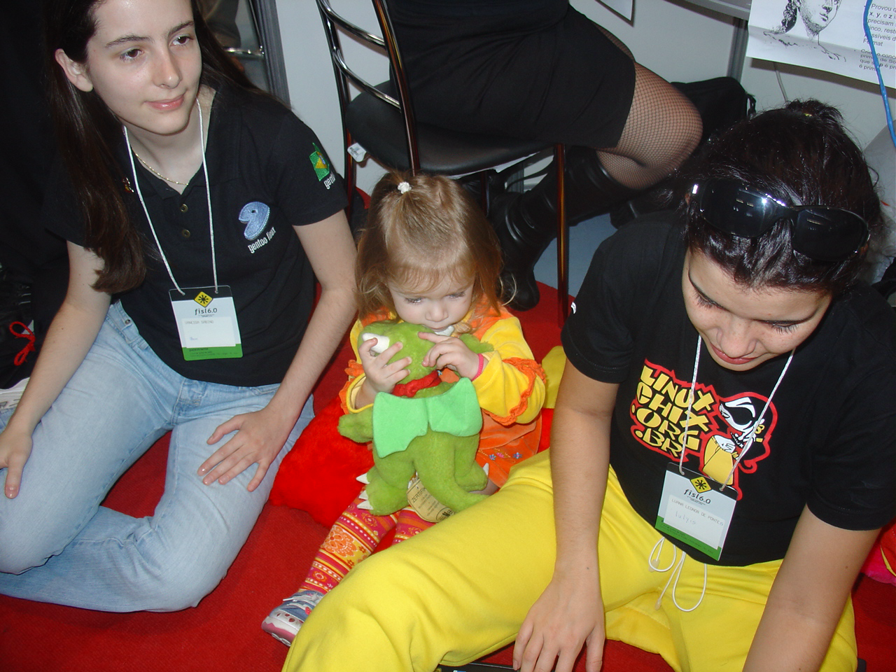 Sentadas num tapete e à frente da Renata (da qual só aparecem as botas), as integrantes das LinuxChix BR: Bani, AliceIGL e Lulyis, em um FISL do início do século