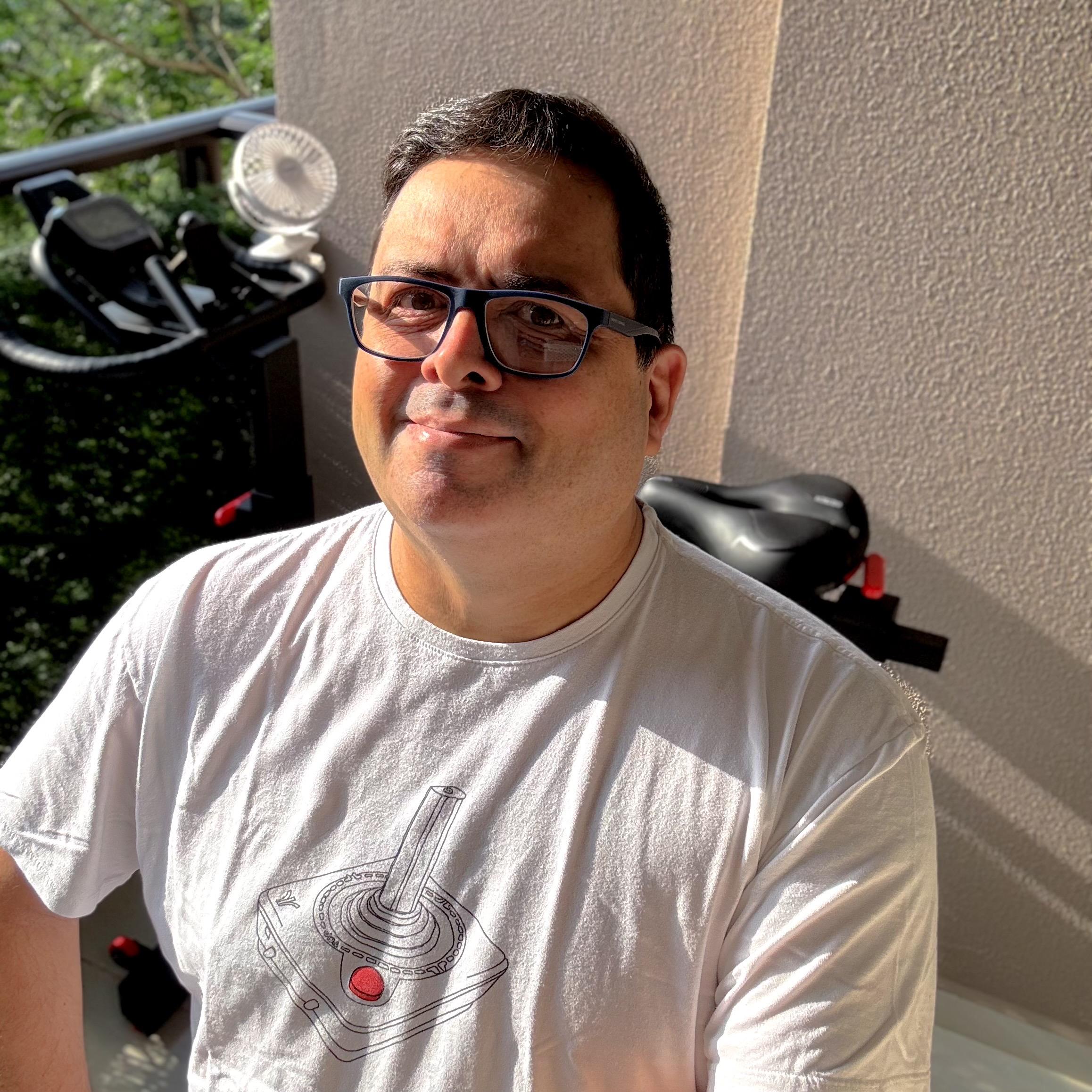 Foto minha olhando para a câmera, com uma camiseta com estampa de joystick de Atari 2600. Ao fundo, a parede ensolarada e uma visão parcial da bicicleta estacionária na minha sacada.