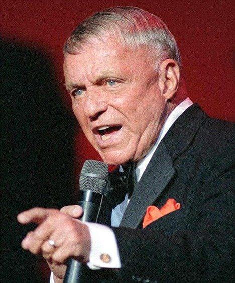 Frank Sinatra, de gravata borboleta e um lenço vermelho no bolso do paletó preto, aponta para a frente, durante seu último show.