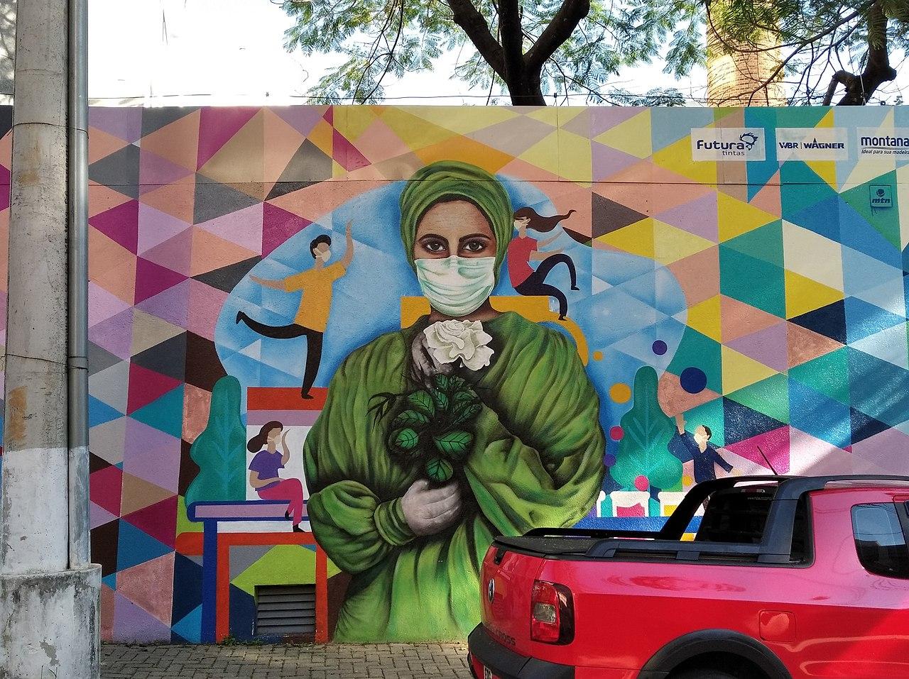 Fotografia do mural em homenagem aos profissionais da saúde, mostrando uma profissional da saúde, paramentada, usando máscara e segurando uma flor branca, durante a pandemia de COVID-19, exposto no Hospital das Clínicas da USP, no prédio do CEAC.