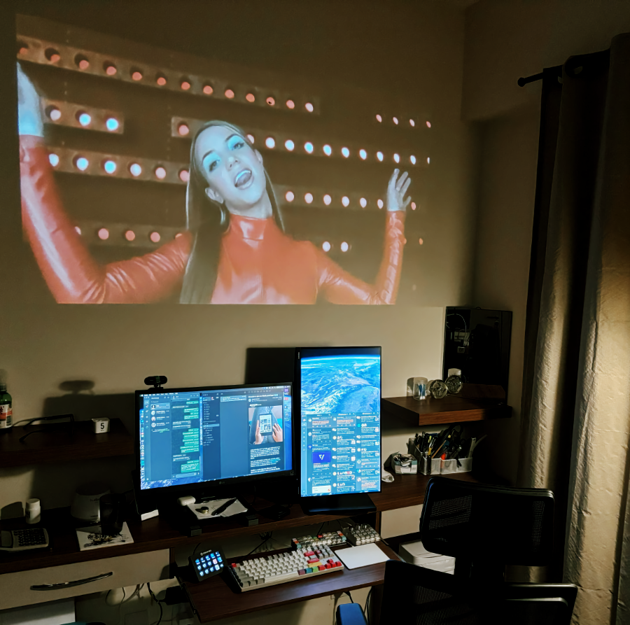 Foto do meu escritório, com uma projeção acima dos 2 monitores. A projeção, feita diretamente na parede, mostra o rosto da cantora Britney Spears, em cena do clip de Oops I Did It Again
