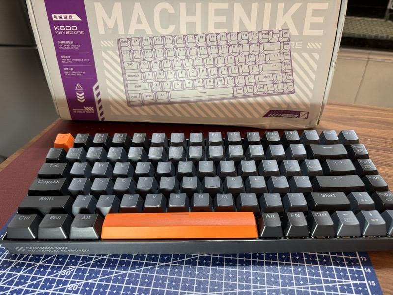 Foto de um teclado Machenike K500, em frente à sua caixa. É um teclado reduzido, com 84 teclas - sem as teclas numéricas adicionais, e com as teclas home, end, page up e page down dispostas na vertical, ao lado direito das teclas enter e backspace