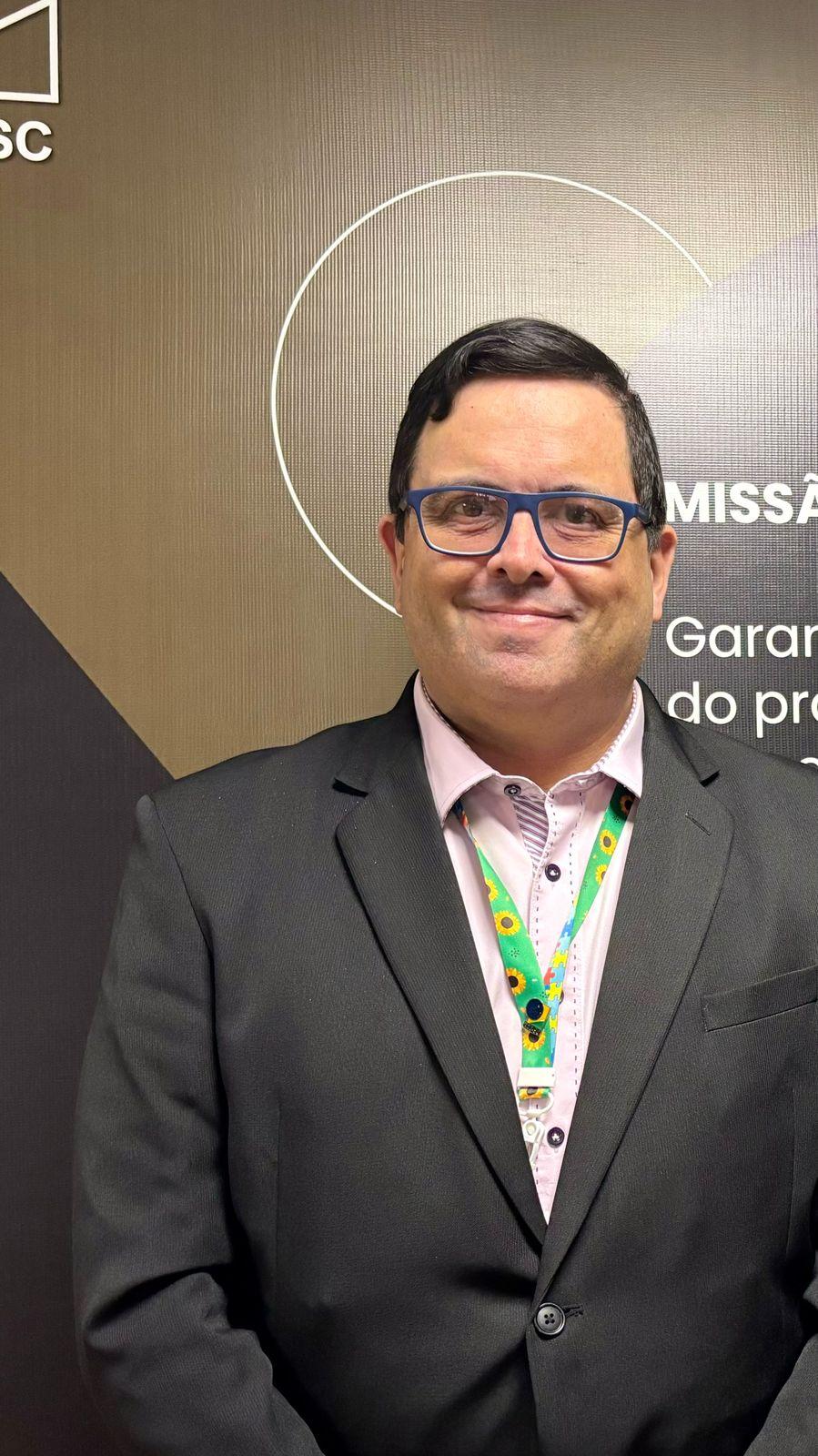 Foto minha em ambiente de trabalho, olhando para a câmera, de paletó preto abotoado e camisa cor de rosa, usando o cordão do girassol, símbolo da presença de deficiência imperceptível (como o TEA, no meu caso)